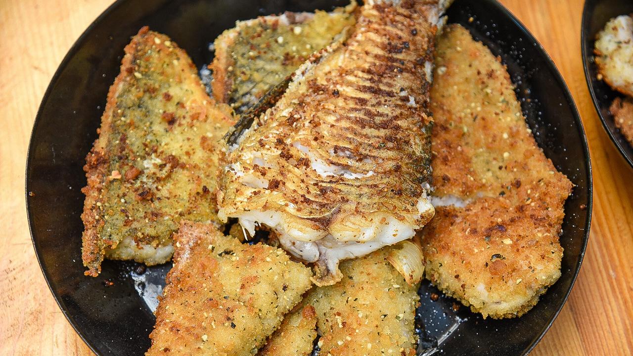 © ORF/Produktion West/Gerhard Mader Mehrere panierte Fischfilets und ein gegrilltes Fischstück auf einem schwarzen Teller.