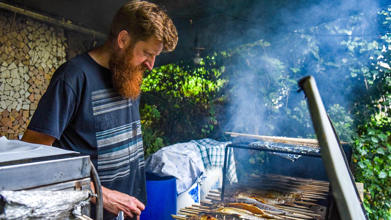 © ORF/Produktion West/Gerhard Mader Franz Wiesmayr steht an einem Grill und überwacht mehrere auf Holzspieße gesteckte Fische (Steckerlfisch) im Freien.