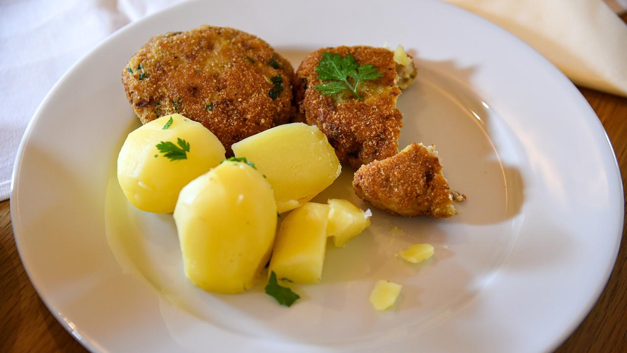 © ORF/Produktion West/Gerhard Mader Zwei goldbraune Fischlaibchen mit Petersilienblatt und Salzkartoffeln auf weißem Teller.