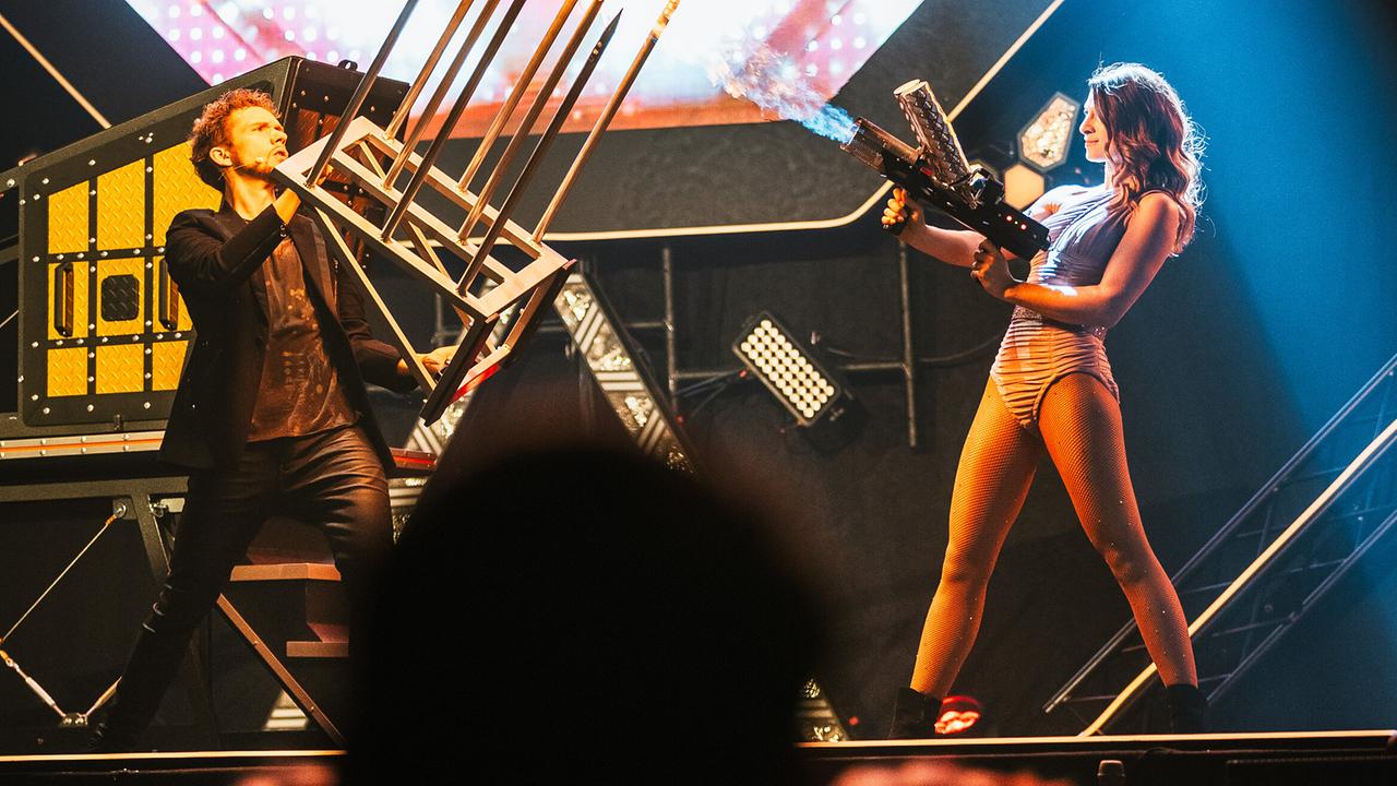 Thommy Ten hält ein Gestell mit brennenden Spitzen und Amélie van Tass steht ihm mit einer Flammenkanone gegenüber.