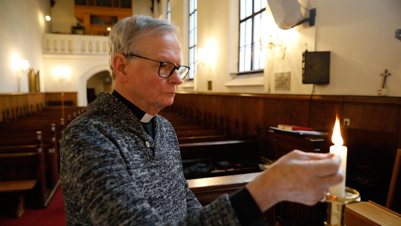 © ORF/Feature Film/Martin Betz In einer Kirche zündet Reverend Canon Patrick Curran vor den leeren Sitzreihen eine Kerze an, während Tageslicht durch große Fenster fällt.