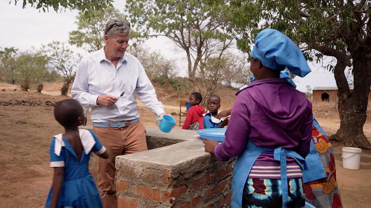 Karl Hohenlohe im Freien mit einer Mitarbeiterin von Marys Meals, die Essen an mehrere Kinder verteilt, an einer einfachen Ausgabestelle in Malawi