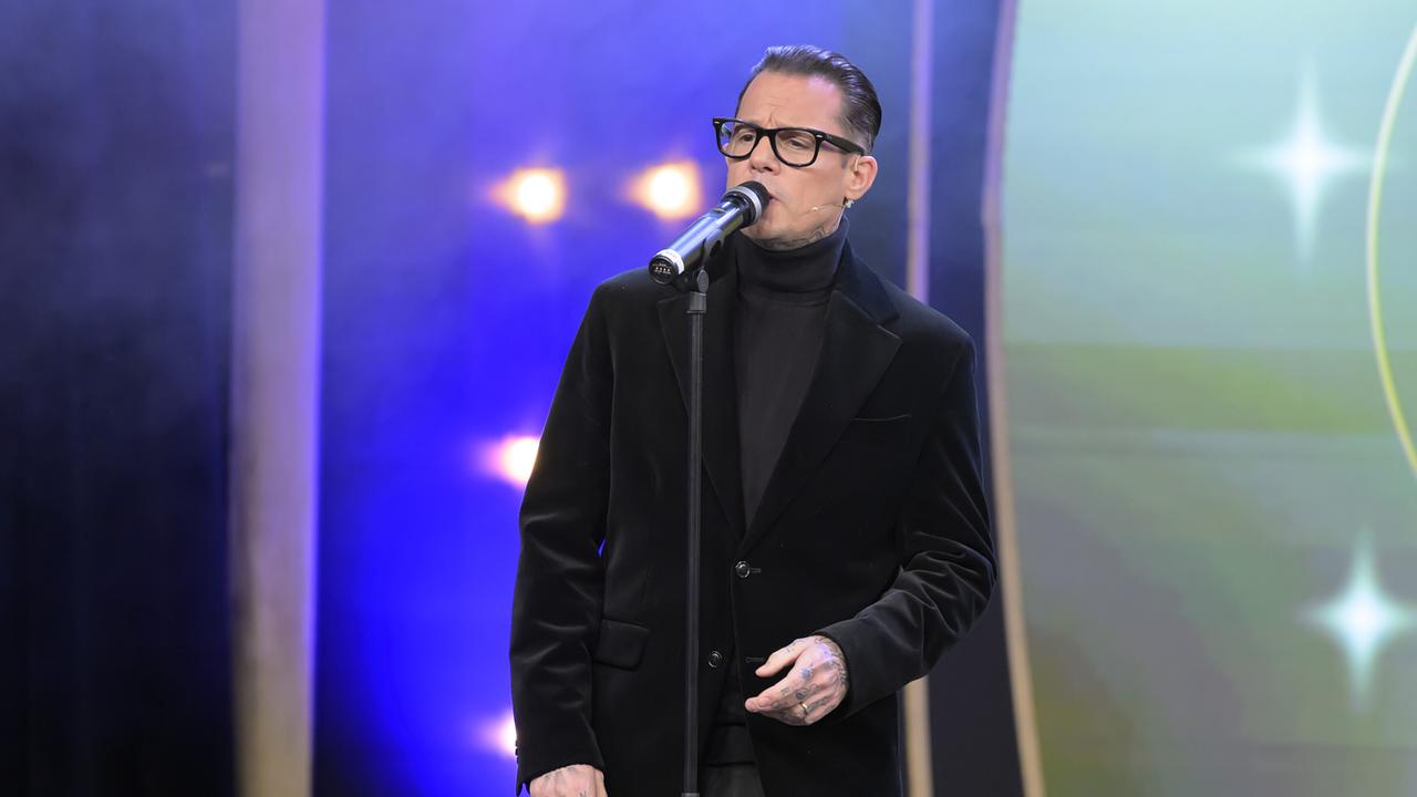 Max Schmiedl  in dunklem Anzug und Rollkragenpullover singt vor einem Mikrofon auf der Bühne.