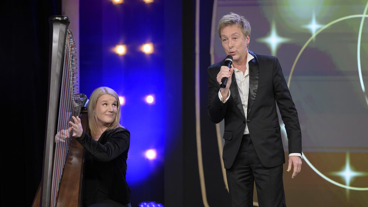 Max Müller im Smoking mit Mikro in der Hand steht neben einer Musikerin mit Harfe auf der Bühne