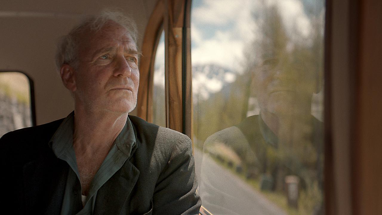 Der alte Andreas Egger sitzt in einem Zug und blickt nachdenklich aus dem Fenster in eine bergige Landschaft.