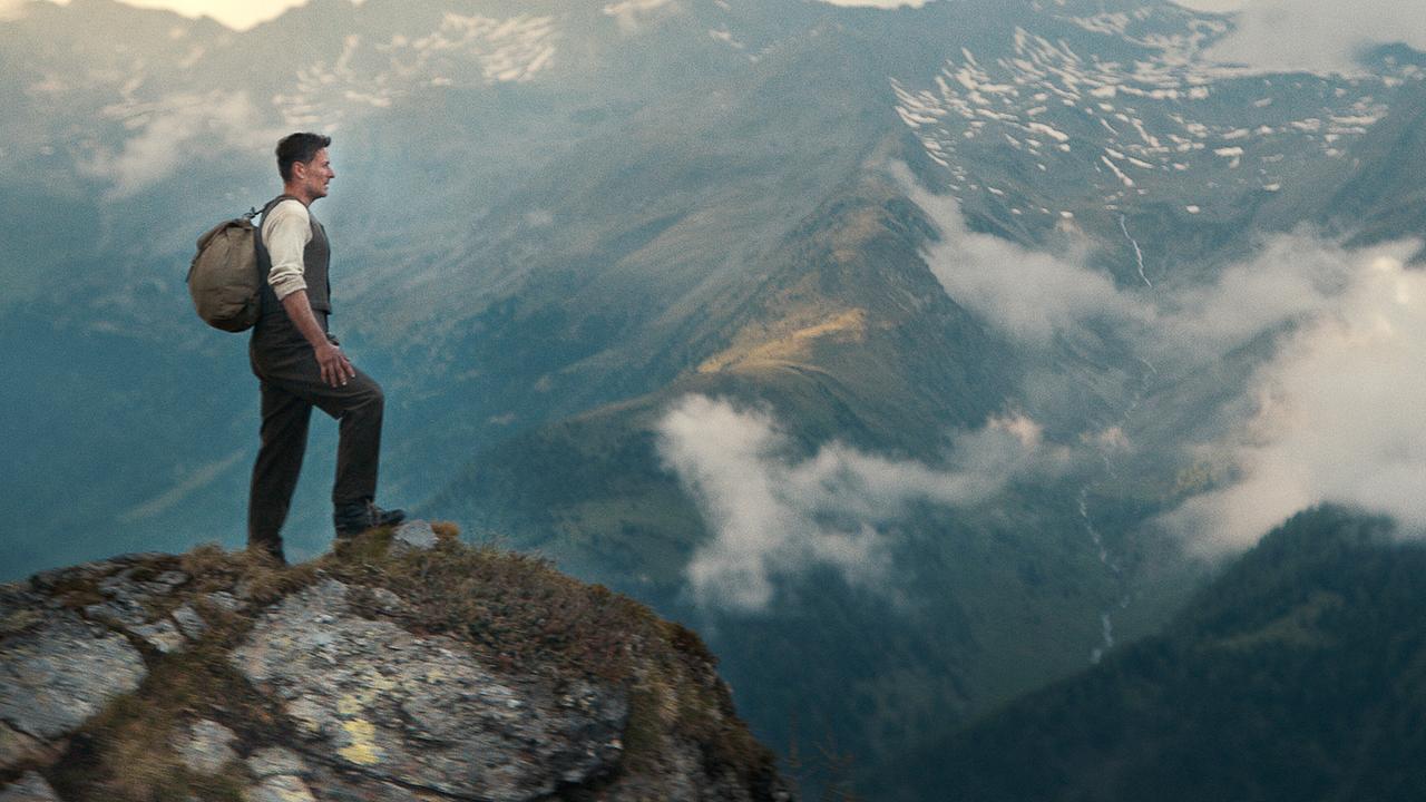 Der junge Andreas Egger steht mit Rucksack auf dem Rücken auf einem Felsen und blickt über eine wolkenverhangene Bergkette.