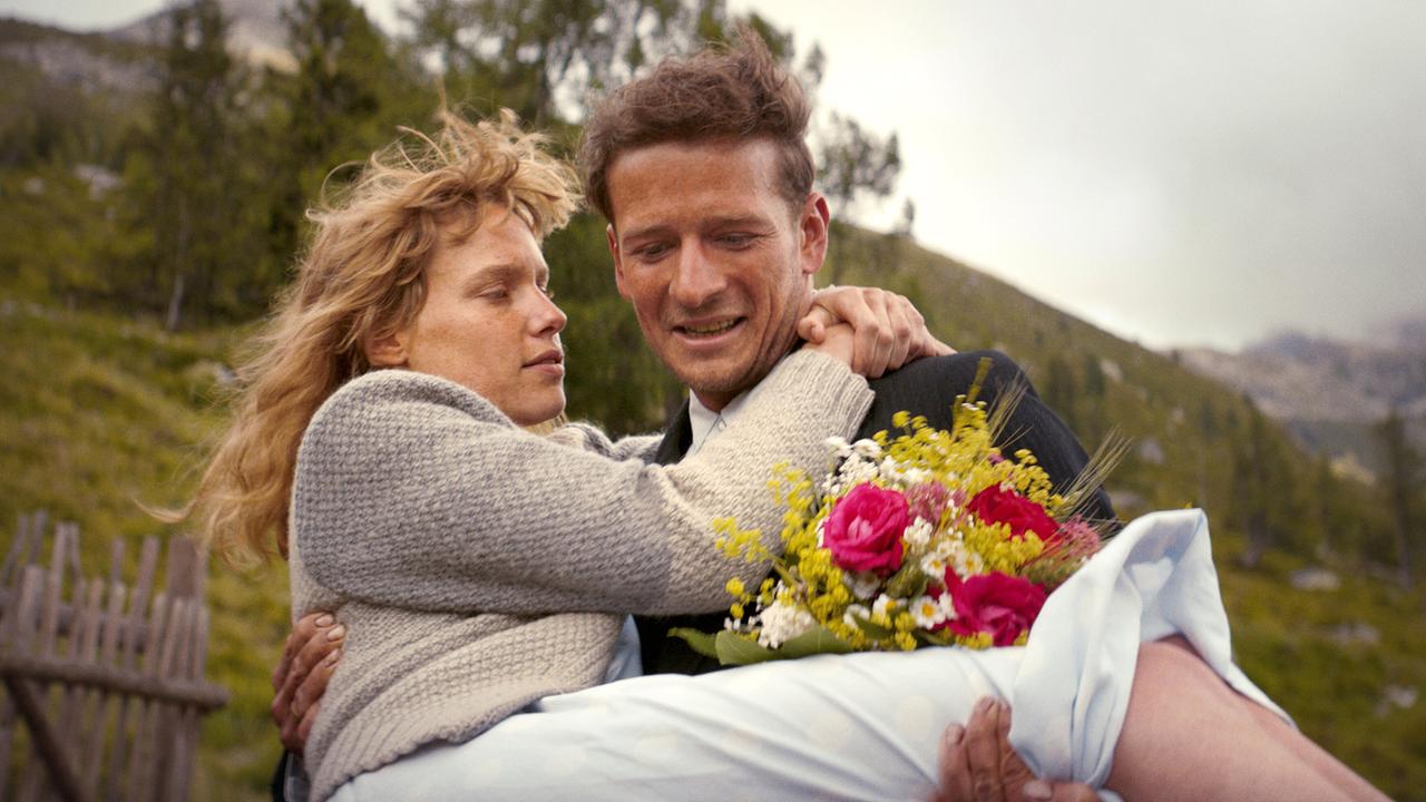 Der junge Andreas Egger trägt Marie in einem weißen Kleid und einen bunten Blumenstrauß in seinen Armen vor einer alpinen Kulisse.