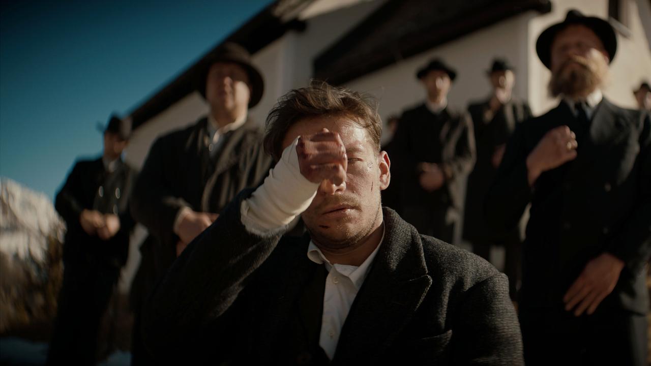 Der junge Andreas Egger sinkt erschöpft zu Boden, hinter ihm eine Gruppe von Männern in traditioneller Kleidung vor einer Bergkapell