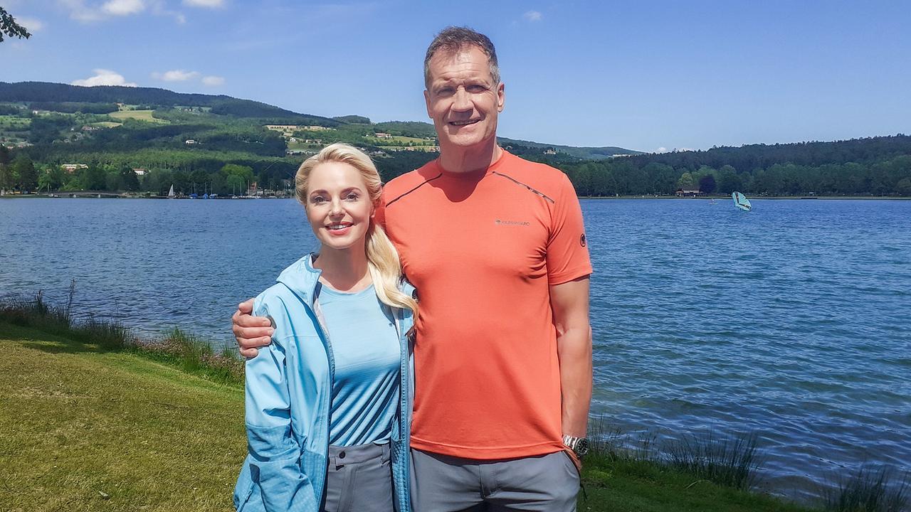 Armin Assinger steht mit Silvia Schneider, um die ihr einen Arm gelegt hat, an einem Seeufer vor einer hügeligen Landschaft bei sonnigem Wetter.