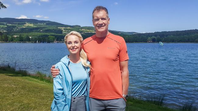 Armin Assinger steht mit Silvia Schneider, um die ihr einen Arm gelegt hat, an einem Seeufer vor einer hügeligen Landschaft bei sonnigem Wetter.