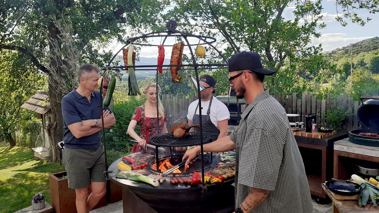 Armin Assinger, Silvia Schneider und Robert Buchberger stehen im Garten rund um eine Grill-Feuerstelle, auf der Nino Sifkovits Fleisch und Gemüse grillt.
