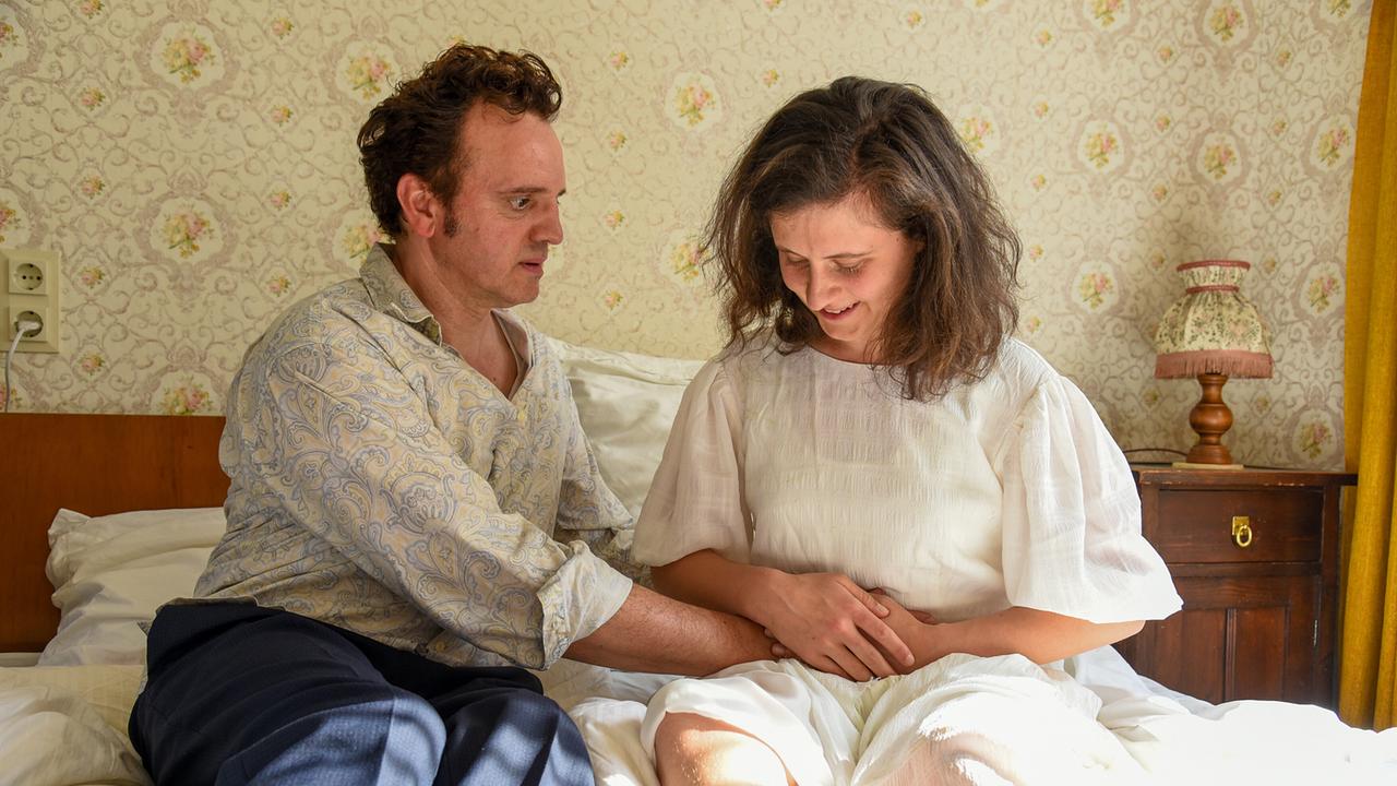 Sepp und Elfi sitzen im Bett, während Sepp liebevoll die Hand auf Elfis Bauch legt.