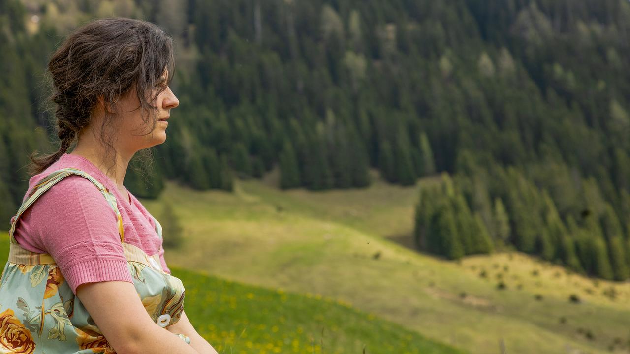 Elfi sitzt auf einer Wiese nahe dem Elternhaus von Sepp und blickt über eine bewaldete Berglandschaft.