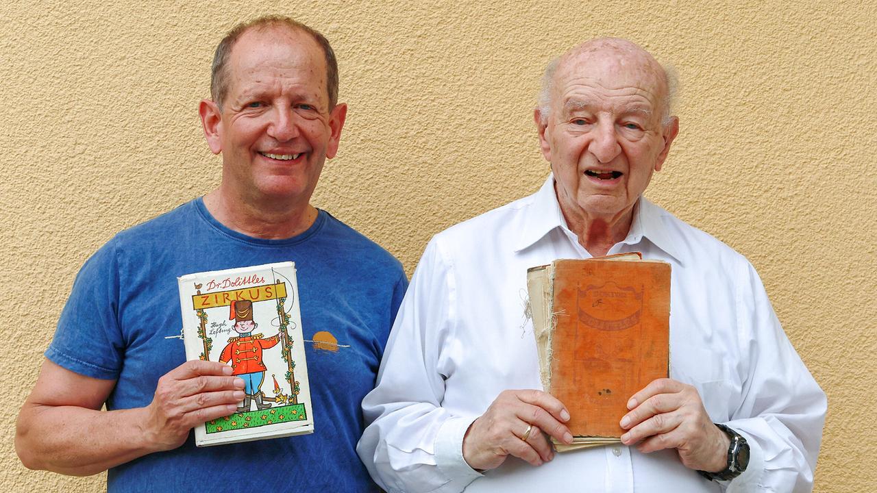 Mario Merl und Vater Harry Merl stehen vor einer hellen beigen Wand und halten jeweils ein Buch in den Händen