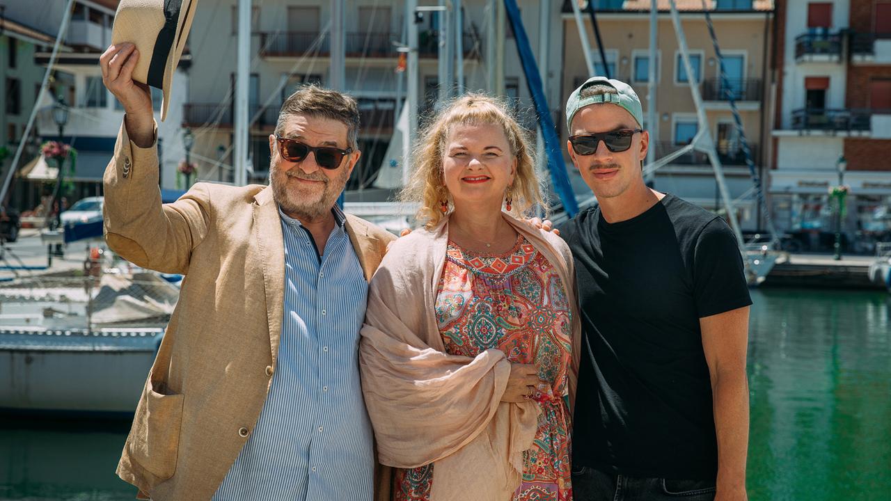 Erwin Steinhauer, Ulrike Beimpold und Matthias Franz Stein posieren gut gelaunt vor Segelbooten in einem sonnigen Hafen.