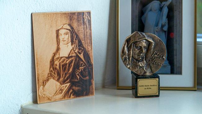 Auf einem Fenstersims lehnt ein Porträt von Edith Stein auf einer Holzplatte und und steht eine Metallskulptur mit ihrem Porträt, im Hintergrund ist ein gerahmtes sakrales Motiv zu sehen.