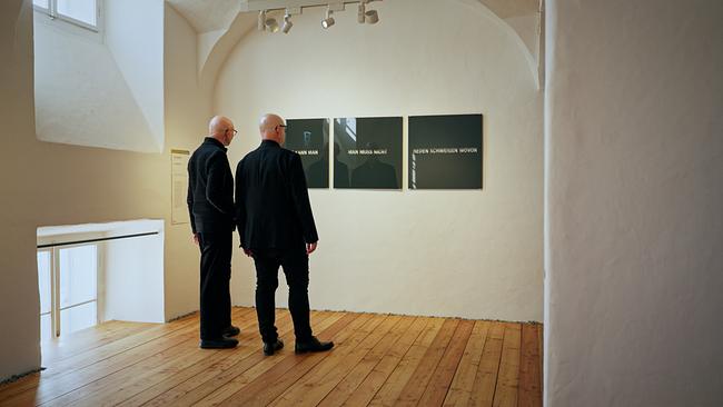 Künstler Leo Zogmayer und Psychiater, Psychologe und Philosoph Michael Lehofer betrachten in einem hellen Ausstellungsraum eine minimalistische Wandarbeit mit mehreren Texttafeln.