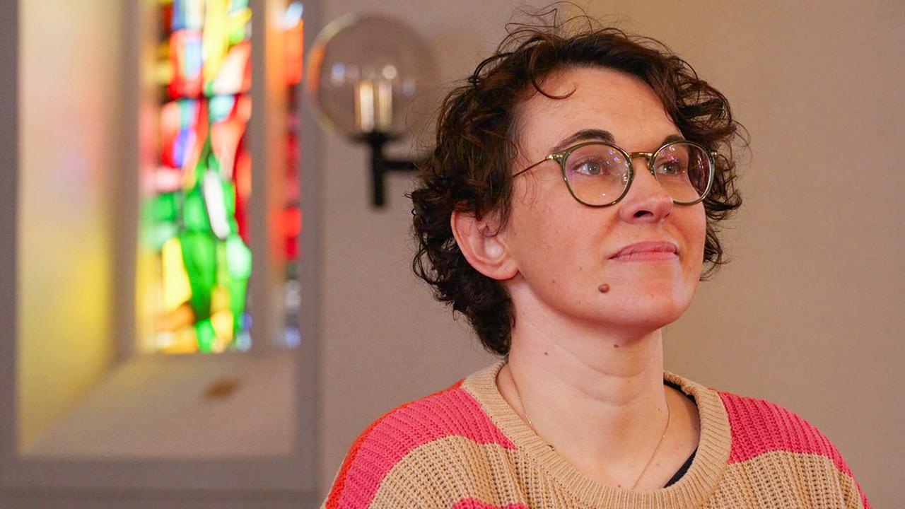Pfarrerin Mag. Iris Haidvogel in einem Innenraum, im Hintergrund ein farbiges Glasfenster und eine Wandlampe.