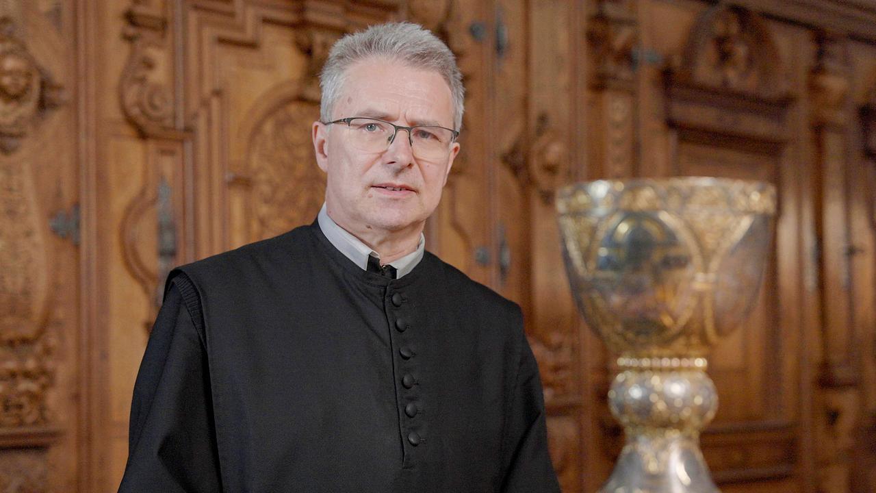 Pater Altman Pötsch mit dem Tassilokelch vor einer reich verzierten Holzwand.