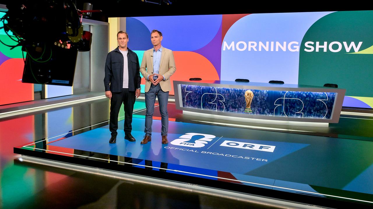 © ORF/Thomas Ramstorfer Michael Liendl und Christian Diendorfer stehen auf der Studiofläche einer farbenfrohen „Morning Show“, umgeben von Kameratechnik und Scheinwerfern, mit dem Moderationstisch und einer goldenen Fußball‑WM-Trophäe im Hintergrund