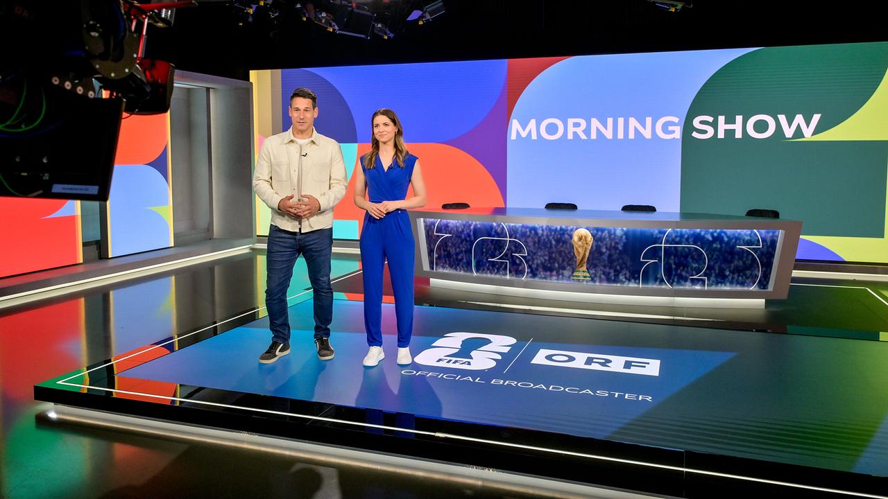 Helge Payer und Kristina Inhof stehen auf der Studiofläche einer farbenfrohen „Morning Show“, umgeben von Kameratechnik und Scheinwerfern, mit dem Moderationstisch und einer goldenen Fußball‑Trophäe im Hintergrund.