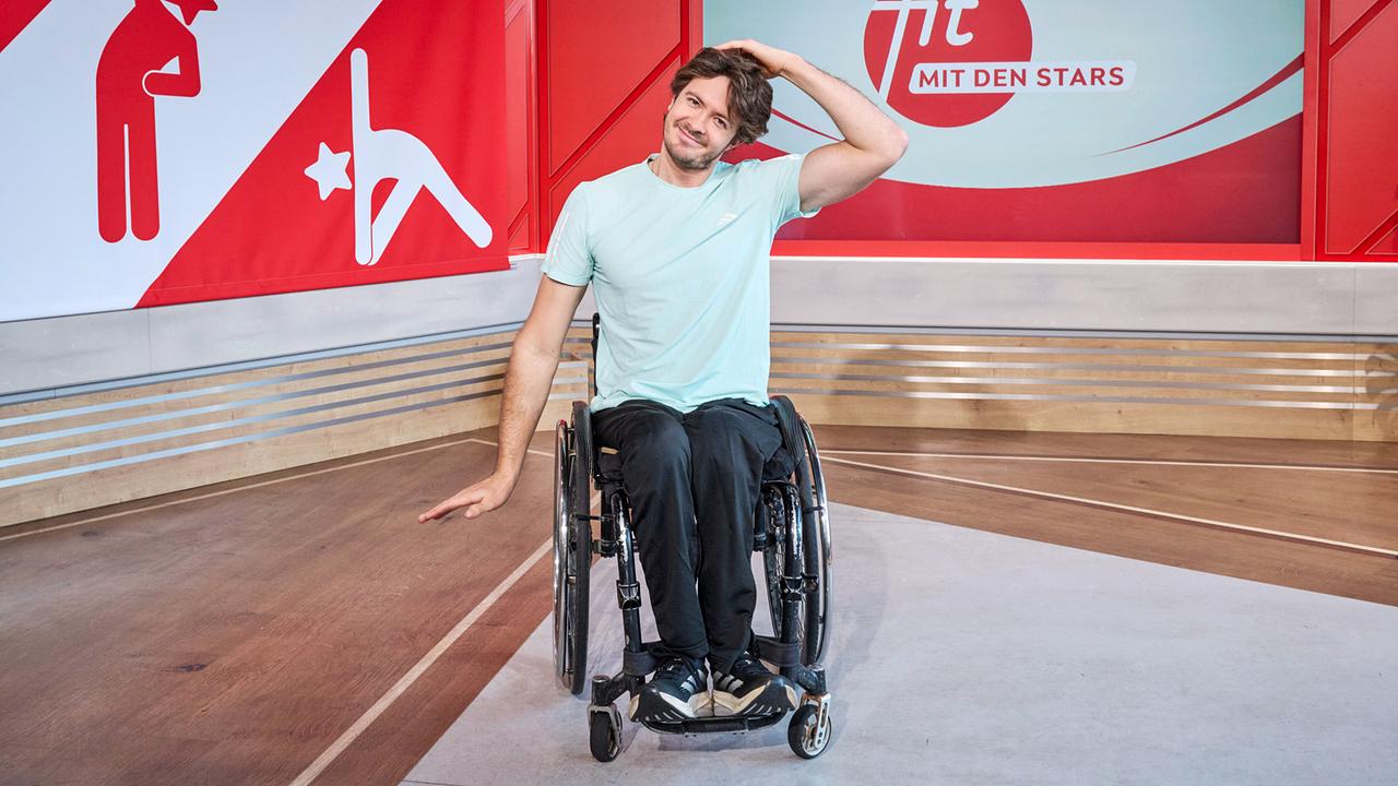 Nico Langmann sitzt in einem Rollstuhl im TV-Studio und führt vor der „Fit mit den Stars“-Kulisse eine Nacken-Dehnübung aus.