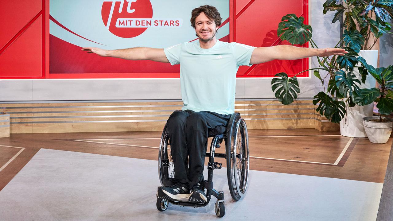 Nico Langmann sitzt im Rollstuhl mit seitlich ausgestreckten Armen im TV‑Studio vor dem Logo „Fit mit den Stars“.