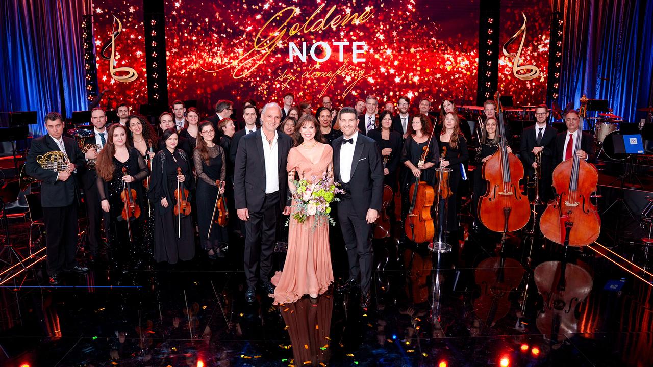 Auf einer festlich beleuchteten Bühne stehen elegant gekleidete Musikerinnen und Musiker mit ihren Instrumenten, vorne begleitet von Martin Gellner, Leona König und Norbert Oberhauser, vor einem großen Hintergrund mit der Aufschrift „Goldene Note“.