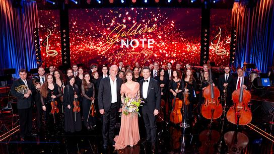 Auf einer festlich beleuchteten Bühne stehen elegant gekleidete Musikerinnen und Musiker mit ihren Instrumenten, vorne begleitet von Martin Gellner, Leona König und Norbert Oberhauser, vor einem großen Hintergrund mit der Aufschrift „Goldene Note“.
