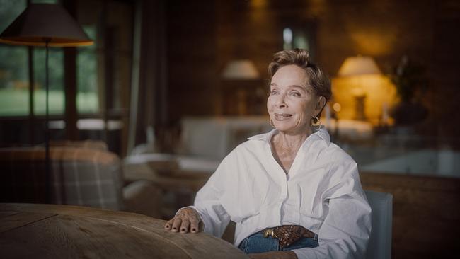 © ORF/FFP Filmproduktion Monika Peitsch in weißer Bluse sitzt in einem warm ausgeleuchteten, stilvollen Innenraum an einem Holztisch.