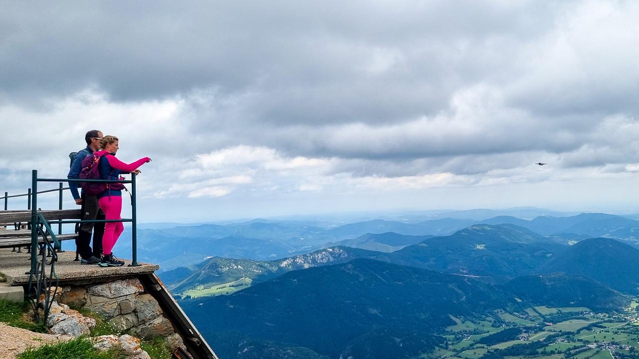 © ORF/Kiwi TV Christine Reiler und Peter Gross auf dem Schneeberg blicken von einer Aussichtsplattform über eine weite Berg- und Tallandschaft.