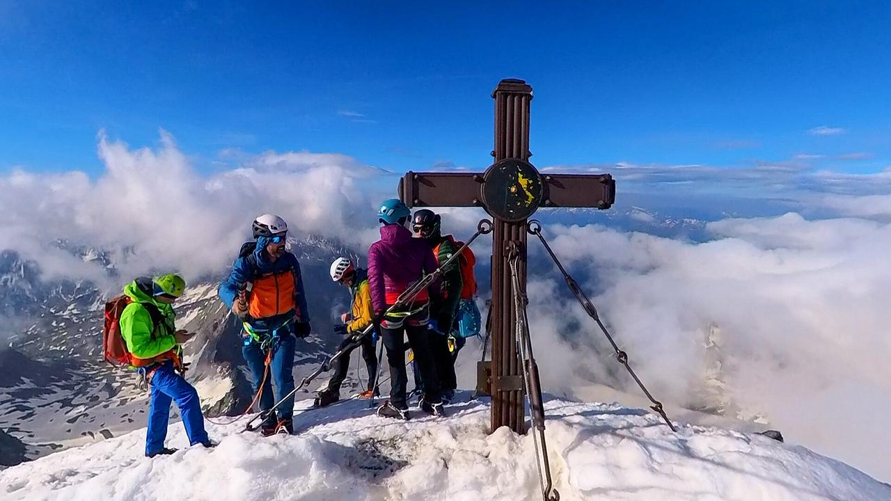 © ORF/Kiwi TV Mehrere Alpinisten stehen auf dem Großglockner auf einem verschneiten Gipfel neben einem großen Gipfelkreuz über den Wolken.