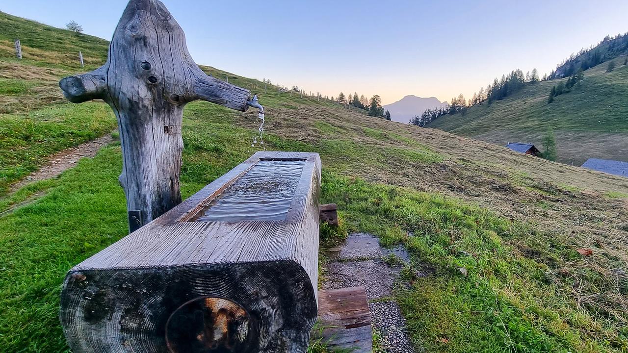 © ORF/Kiwi TV Ein rustikaler Holzbrunnen steht auf der Almwiese bei der Erlbachhütte, im Hintergrund liegen verstreut mehrere Hütten.