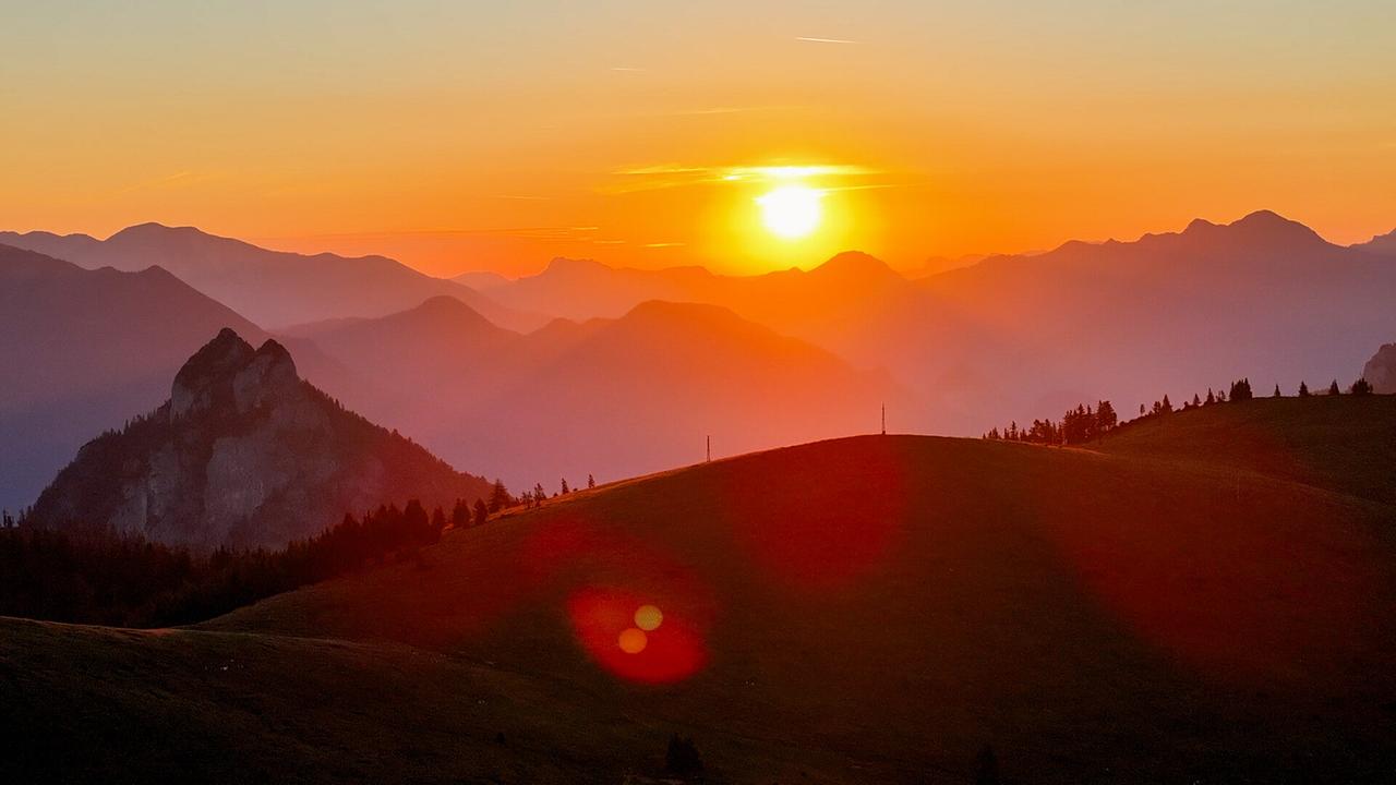 © ORF/Kiwi TV Die Sonne geht glühend orange über einer Berglandschaft mit sanften Hügeln und dunklen Silhouetten auf oder unter.