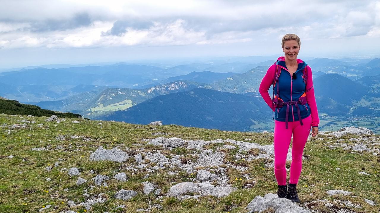 © ORF/Kiwi TV Christine Reiler in pink-blauer Bergsportkleidung steht auf dem Gipfelplateau des Schneebergs mit weitem Talblick.