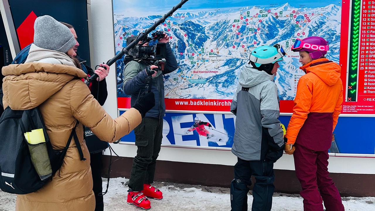 © ORF/Kiwi TV Ein Kamerateam filmt Christine Reiler und Gottfried Bistumer, beide in Skibekleidung, vor einem großen Pistenplan in einem Skigebiet.
