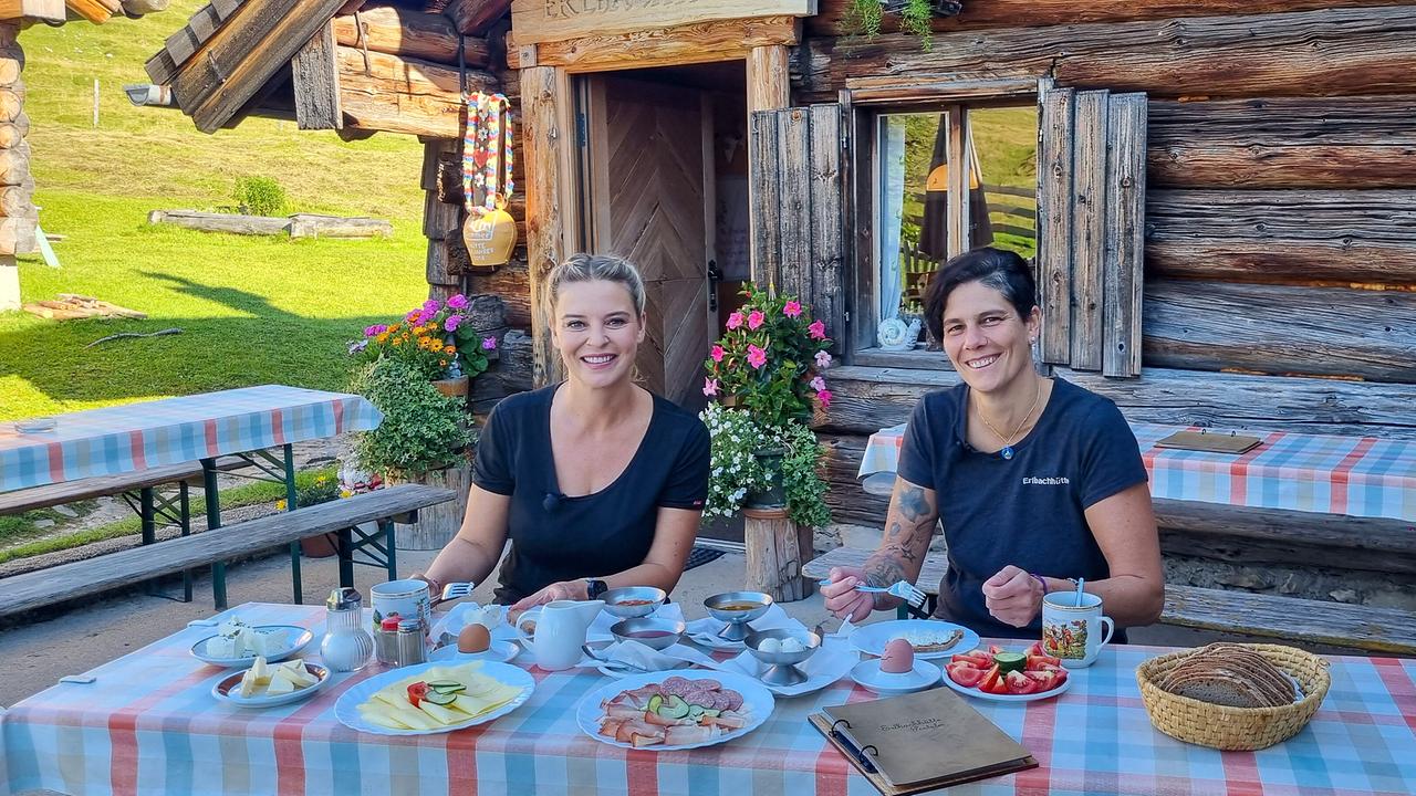 © ORF/Kiwi TV Christine Reiler zu Gast bei der Sennerin Bernadette Gerauer auf der Erlbachhütte. Sie sitzen vor der Almhütte an einem gedeckten Tisch und genießen ein Frühstück im Freien.