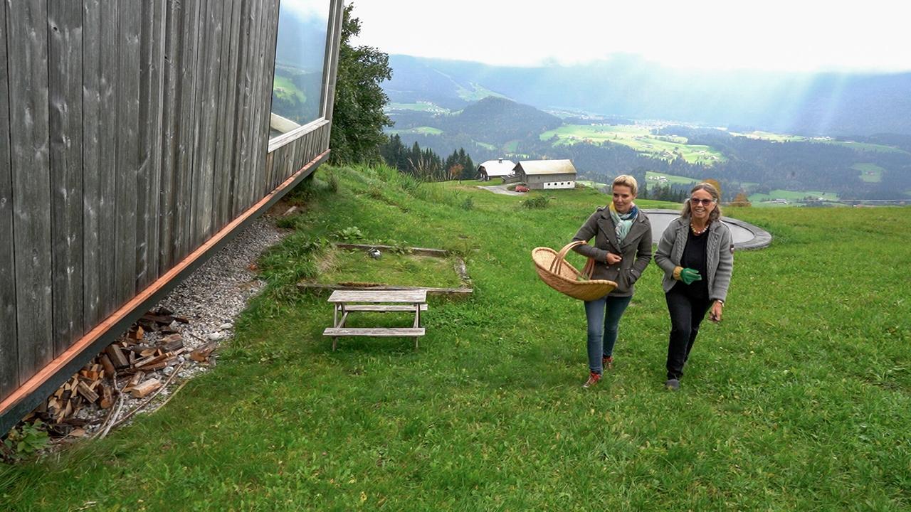 © ORF/Kiwi TV Renee Schröder und Christine Reiler gehen beim Leierhof mit einem Korb neben einem Holzhaus einen steilen Wiesenhang hinauf.