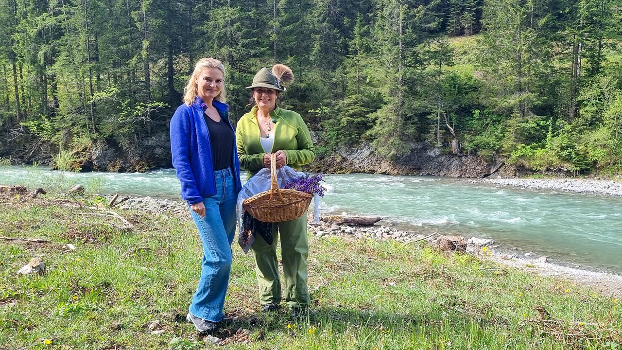© ORF/Kiwi TV Christine Reiler steht lächelnd mit Verena Lämmle, die einen Kräuterkorb in der Hand hält, am Ufer eines wilden Gebirgsflusses.