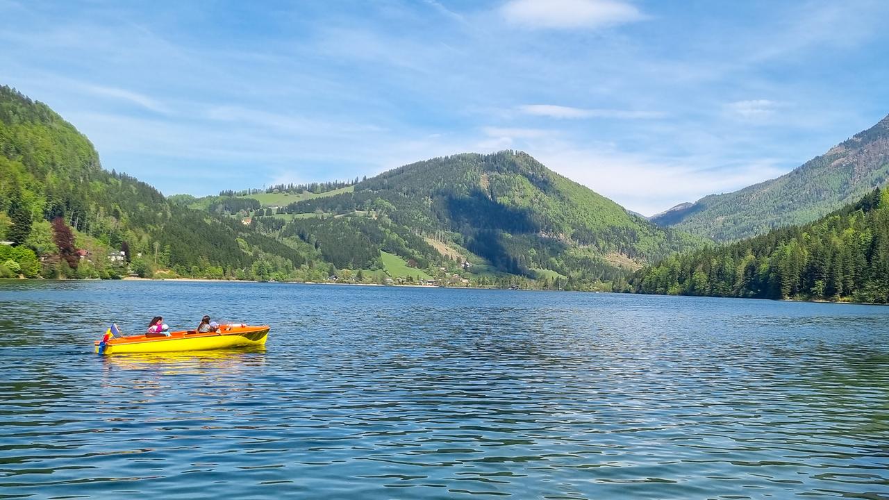 © ORF/Kiwi TV Ein gelbes Boot fährt mit zwei Personen über den WasserCluster Lunz vor grünen Hängen und Wäldern.