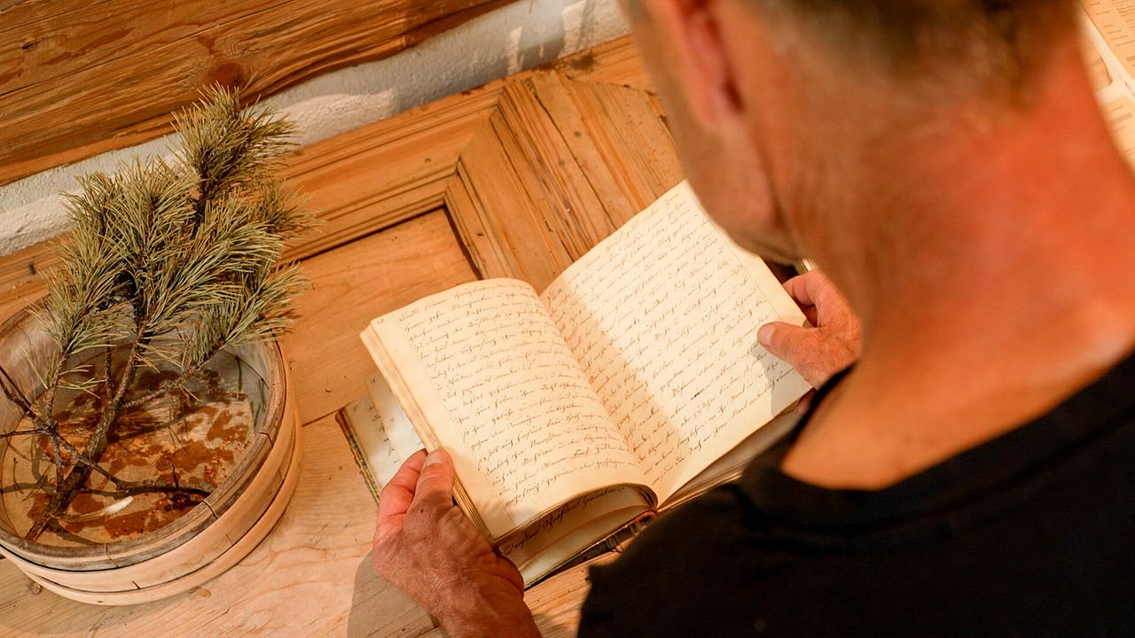 © ORF/Kiwi TV Ein Mann blättert in einem handgeschriebenen alten Buch, einer Sammlung alter Hausmittel, in einer rustikalen Stube.