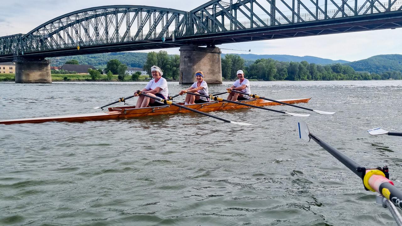 © ORF/Kiwi TV Drei Ruderinnen und Ruderer vom Steiner Ruderclub sitzen unter einer großen Stahlbrücke in einem Holzboot auf einem breiten Fluss.