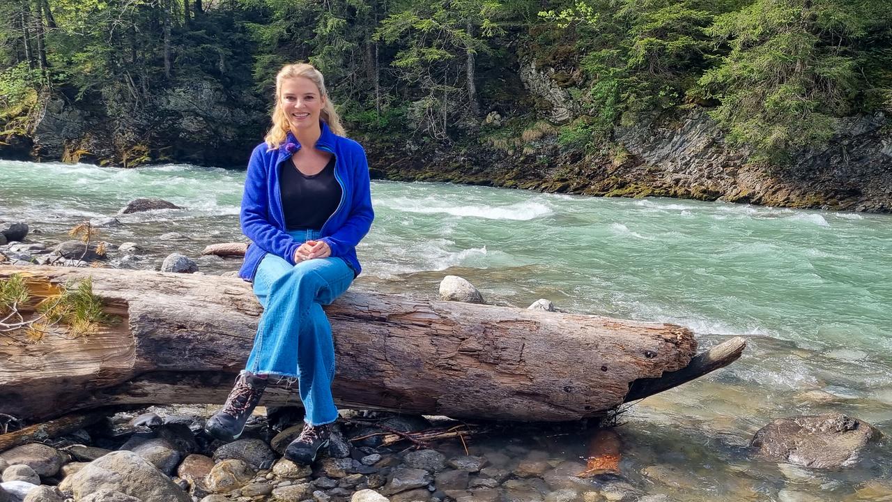 © ORF/Kiwi TV Christine Reiler sitzt auf einem Baumstamm am Ufer an der Lech.