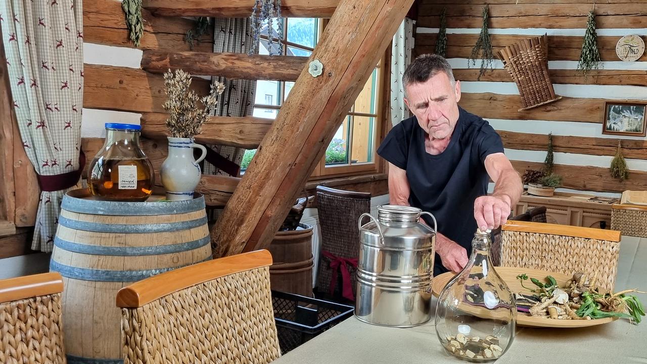 © ORF/Kiwi TV Josef Wildanger hantiert in einer urigen Holzhütte mit Flaschen, Kräutern und Gefäßen auf einem Tisch.