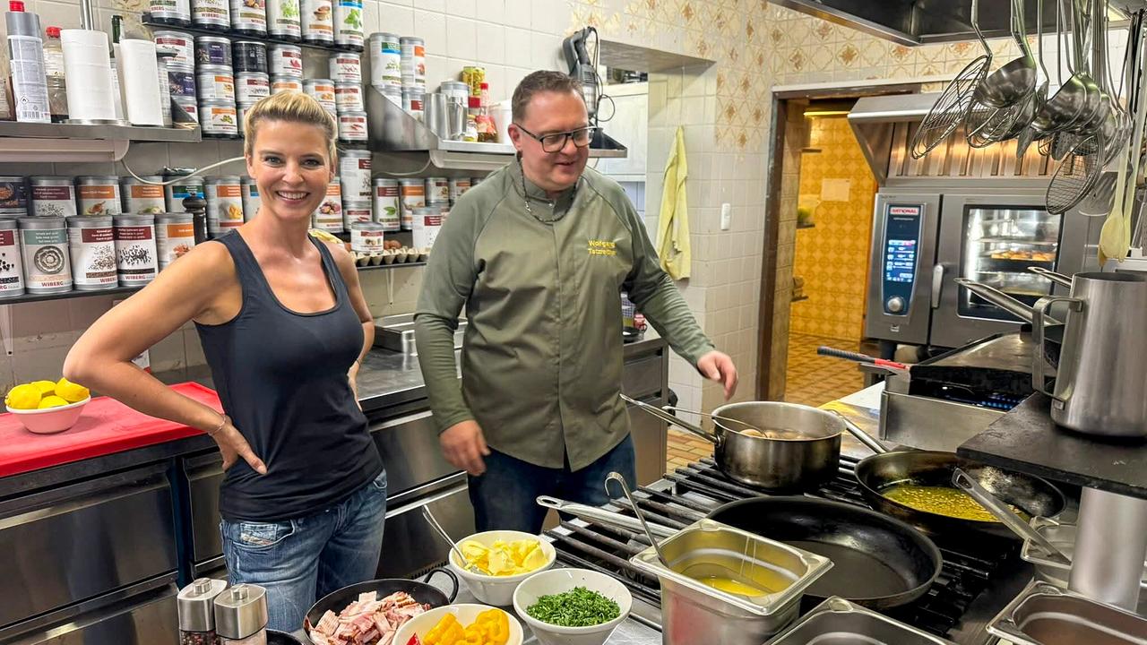 © ORF/Kiwi TV Christine Reiler beim Bruckwirt steht mit einem Koch in einer Profiküche vor vorbereiteten Zutaten und Töpfen.