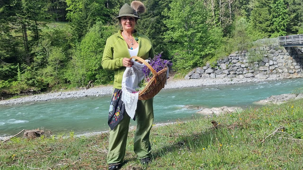 © ORF/Kiwi TV Verena Lämmle in grüner Tracht steht mit einem Korb voller Kräuter und Blumen an einem türkisfarbenen Fluss.