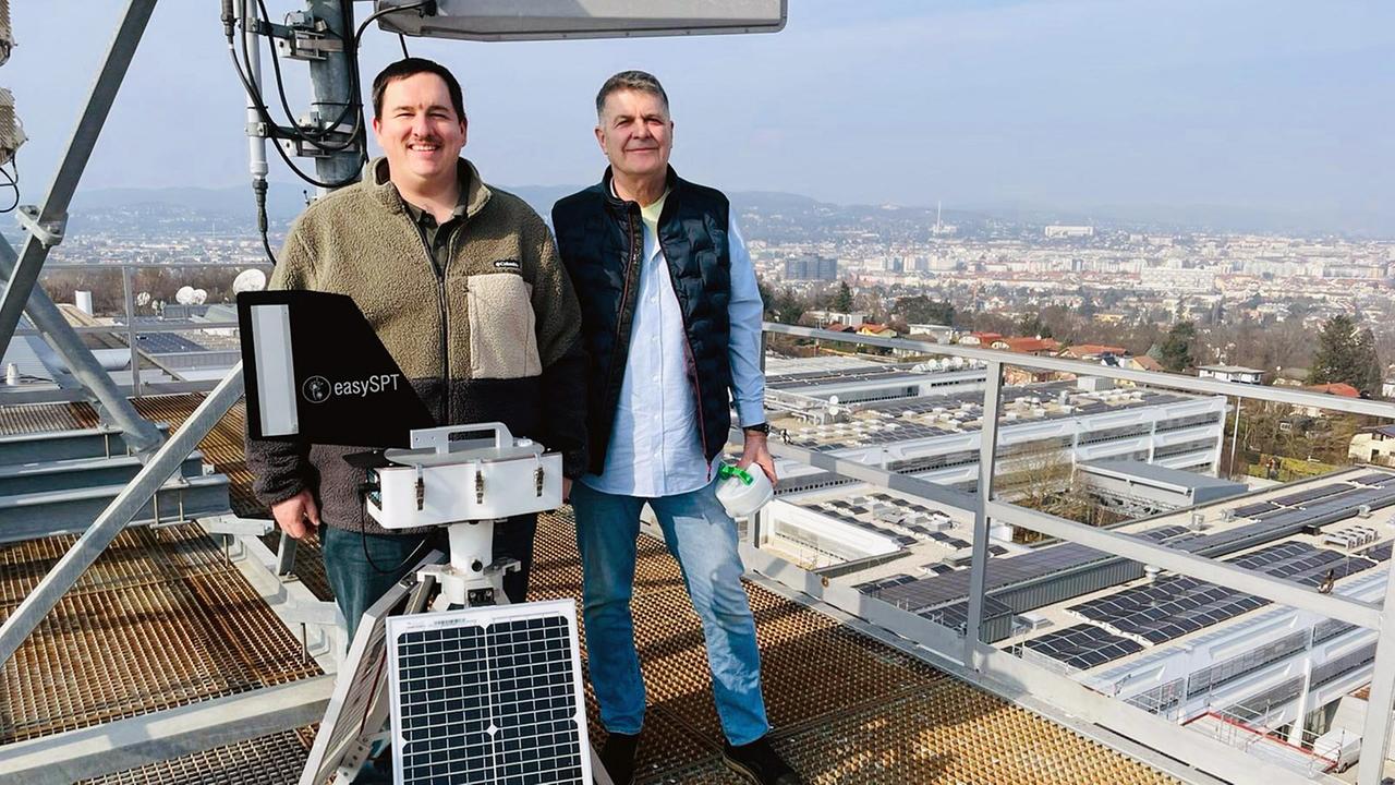 Zwei Personen stehen bei Sonnenschein auf einer Dachplattform des ORF-Zentrums neben einer technischen Messanlage mit Solarpanel und Stadtblick im Hintergrund.