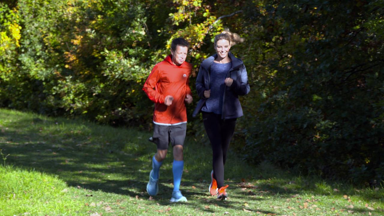 Helmut Steigersdorfer und Christine Reiler joggen gemeinsam auf einem schmalen Waldweg durch grünes und herbstliches Laub.