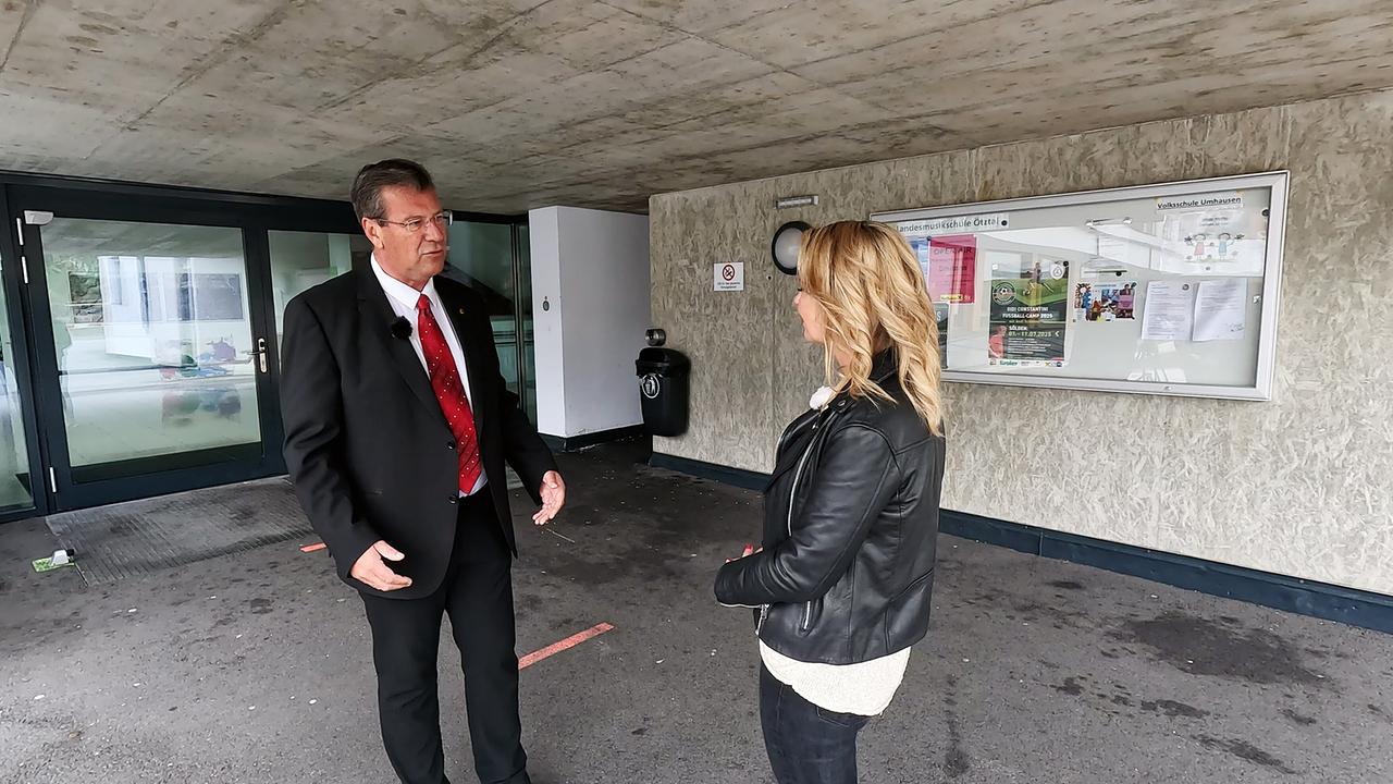 Dr. Christine Reiler im Gespräch mit Jakob Wolf, Bürgermeister von Umhausen vor dem Eingang eines öffentlichen Gebäudes mit Aushangtafeln an der Wand.