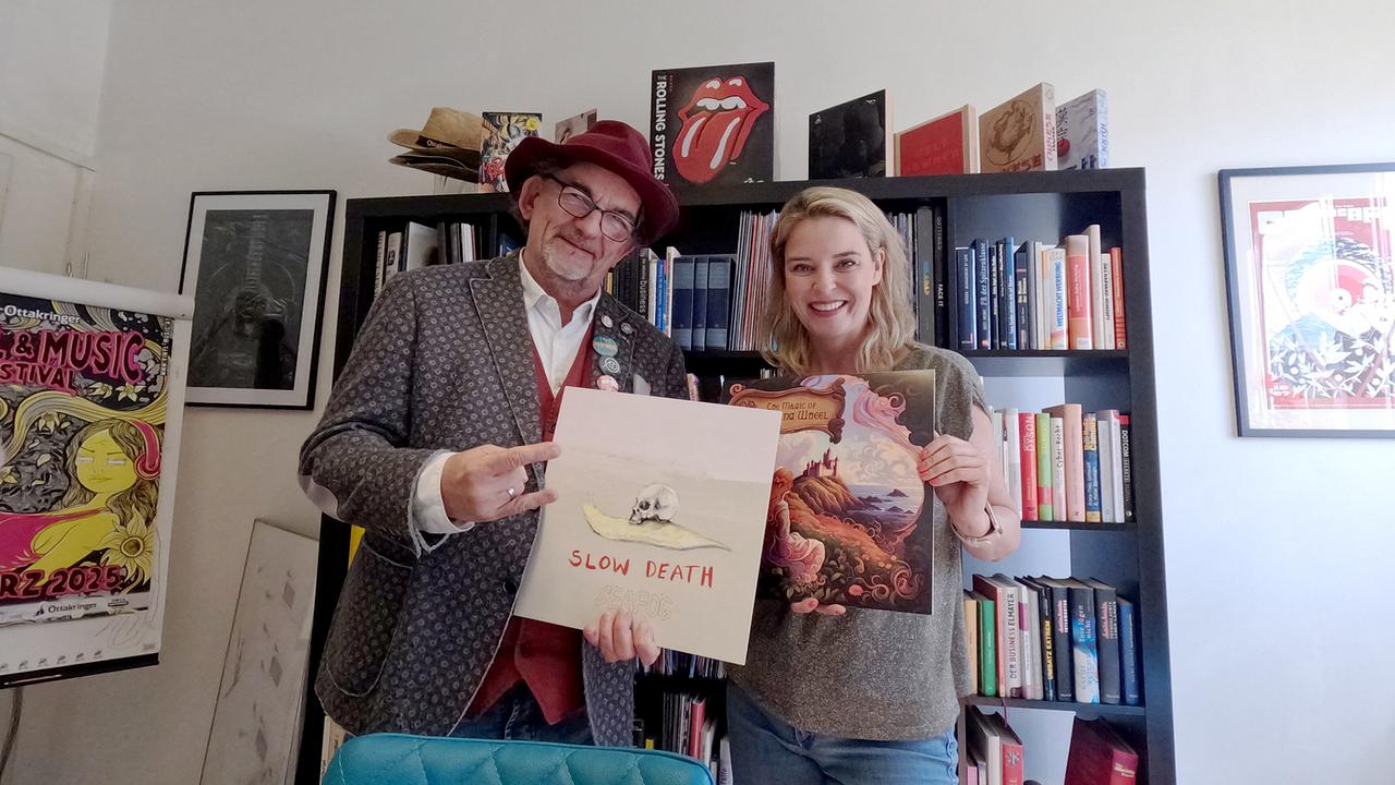 Till Philippi und Dr. Christine Reiler stehen in einem hellen Zimmer und halten Schallplatten in der Hand vor einem schwarzen Regal voller Bücher und Musik.